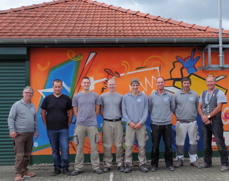 Neun Mitarbeiter der Wittmann GmbH stehen nebeneinander vor einer bunt bemalten Wand des Betriebsgebäudes